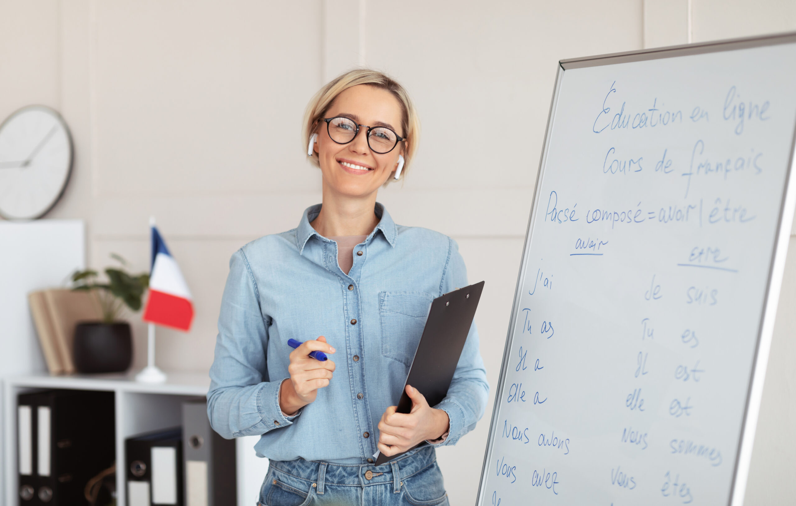 Friendly young female tutor standing near blackboard with French grammar rules, giving web lesson from home. Foreign language professor teaching students online. Remote education concept